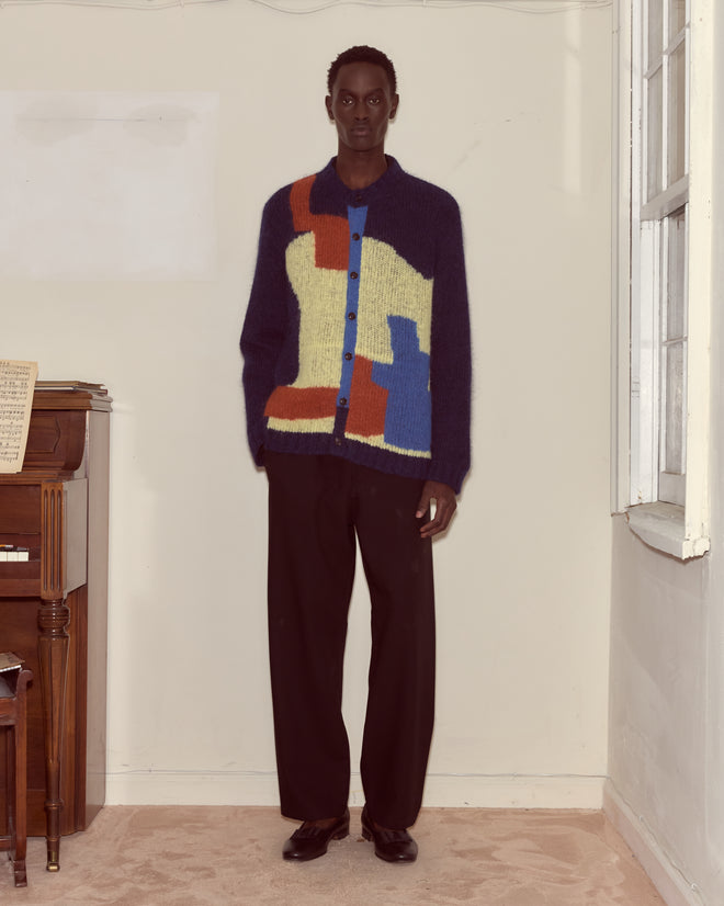 A person stands indoors by a piano and window, wearing the Chapeau Cardigan with colorful abstract colorblocking, paired with black pants and black shoes.