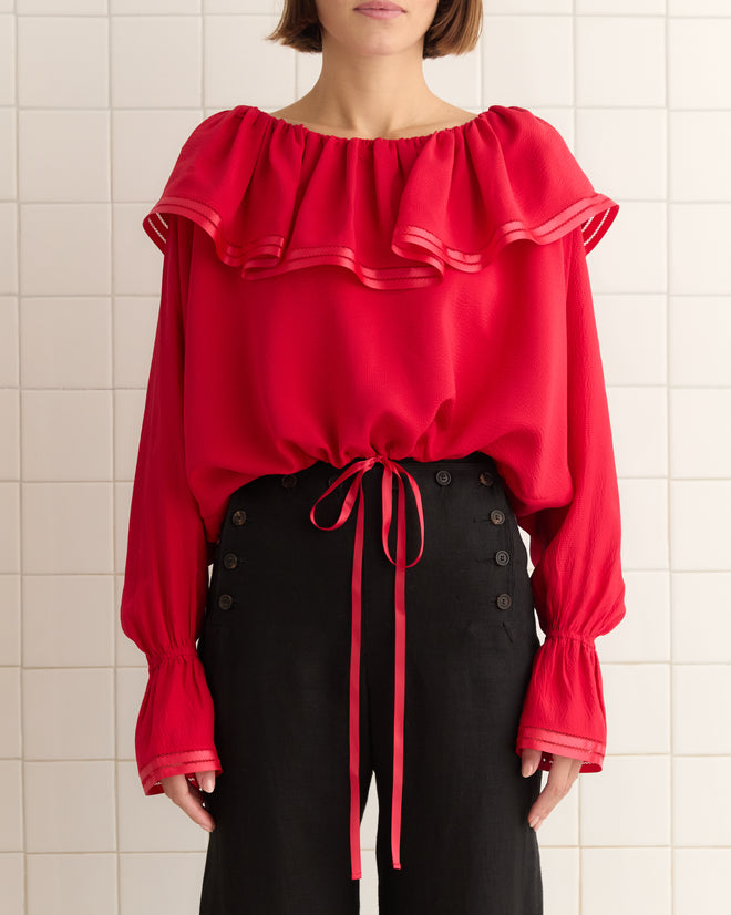 A person wearing a Satin Claret Blouse - Red with a ruffled collar and black buttoned trousers stands against a tiled wall.
