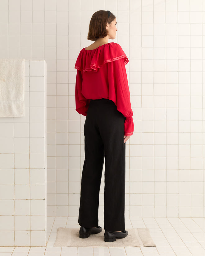 A woman in a Satin Claret Blouse - Red and black pants stands in a tiled room, facing away. Her blouse features a ruffled collar that elegantly enhances her silhouette.