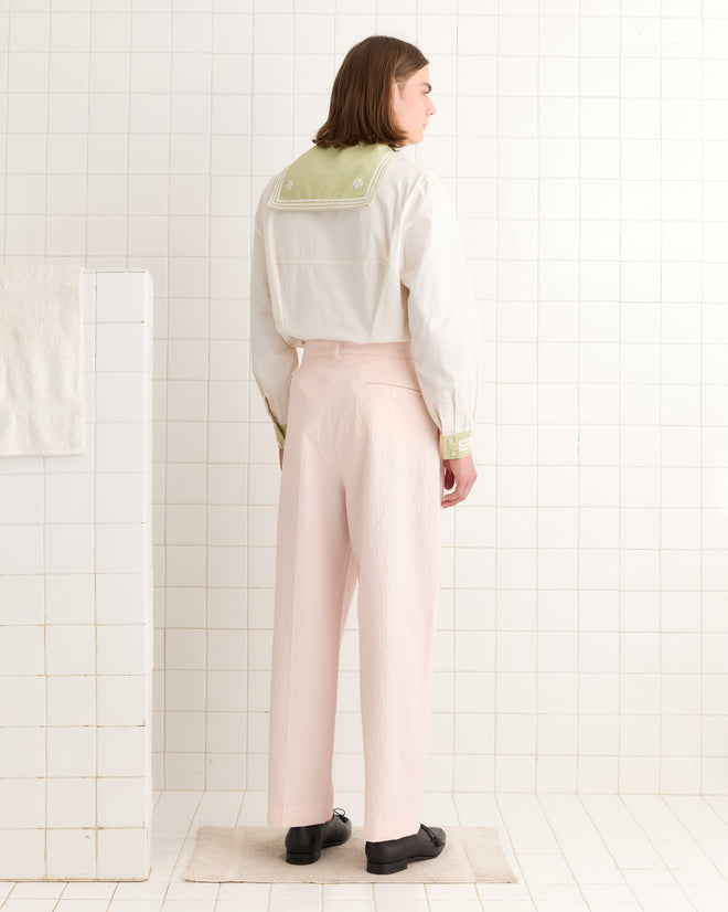 In a white tiled room, a person stands out wearing Club Stripe McNab Trousers in Pink/White, paired with a white shirt subtly detailed with green on the back.