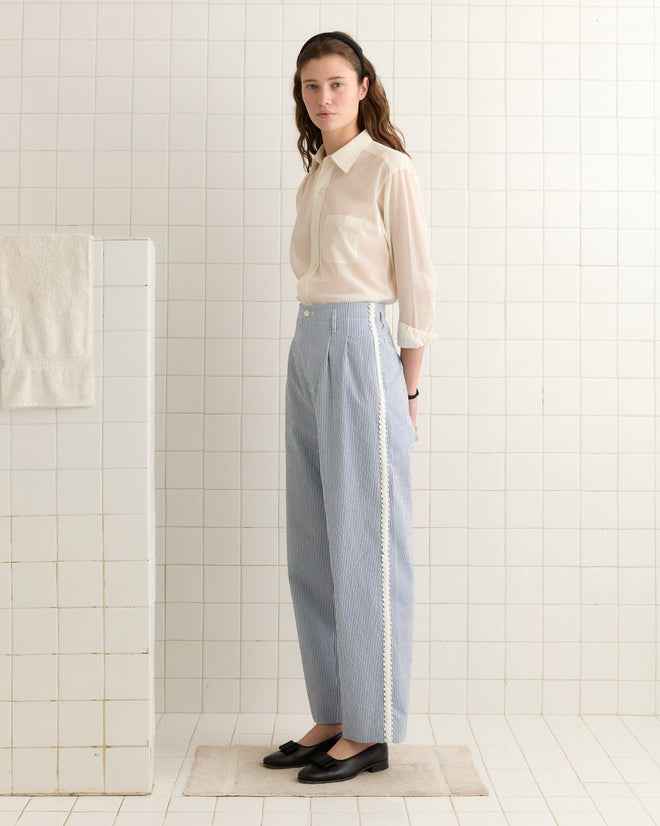 A woman stands in a tiled room wearing a sheer white blouse with ivory trim, Club Stripe Murray Trousers in blue/white, and black shoes, evoking late 20th-century style.