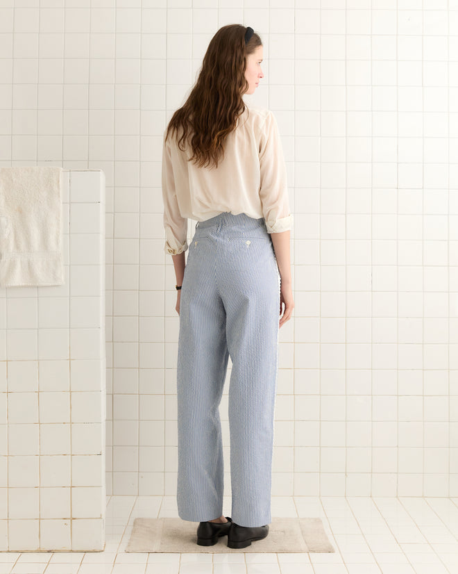 A woman with long hair stands on a rug in a tiled room, facing away, wearing a light blouse with ivory trim and Club Stripe Murray Trousers in Blue/White—evoking late 20th-century style.
