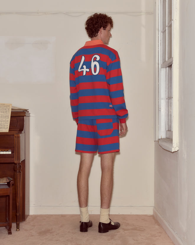 A man in the Clubhouse Rugby Shirt, featuring wide red and blue stripes and the number 46, stands facing a wall near a window.