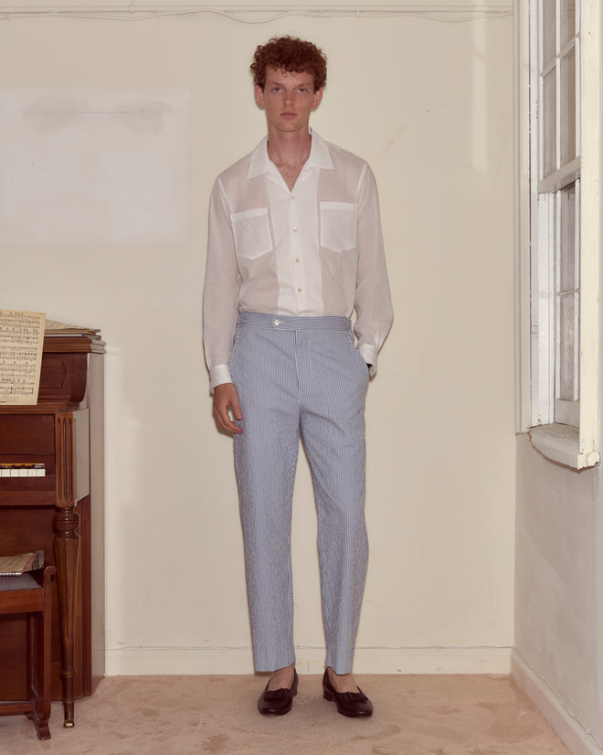 A man stands indoors by a piano, wearing a white shirt and Club Stripe Side-Buckle Trousers in blue/white, finished with dark shoes.