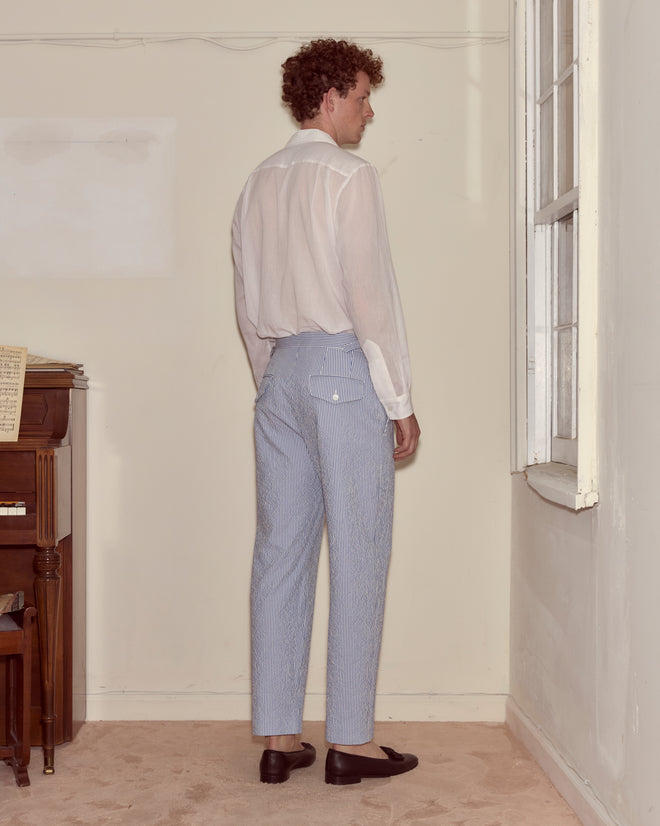 A man in a white shirt and Club Stripe Side-Buckle Trousers - Blue/White stands facing a wall near a window and piano.