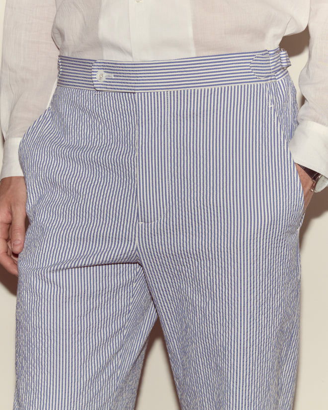 Person wearing a white shirt and Club Stripe Side-Buckle Trousers in blue/white, featuring a relaxed fit with one hand in their pocket.