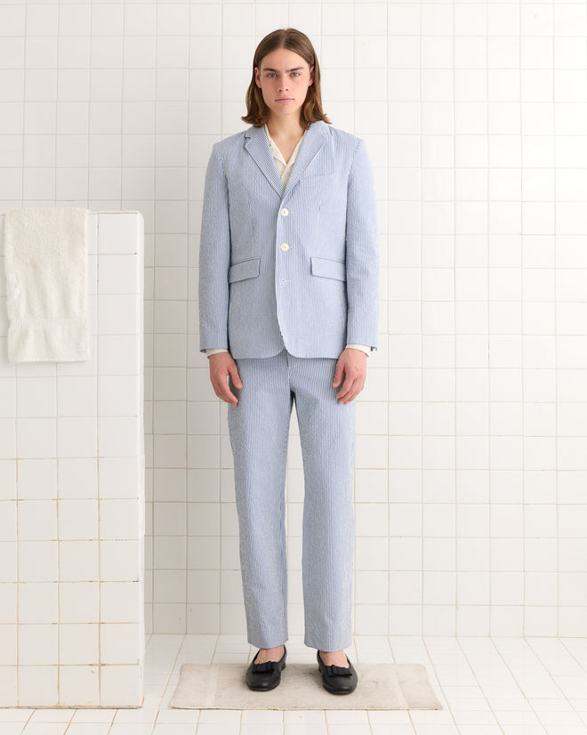 A person in a Club Stripe Single-Breasted Suit Jacket - Blue/White stands in a tiled room, exuding classic elegance, next to a neatly folded towel.