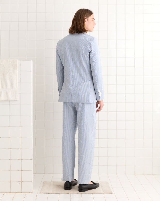 A person wearing a Club Stripe Single-Breasted Suit Jacket in blue/white stands on a small rug in a white-tiled room, facing away.