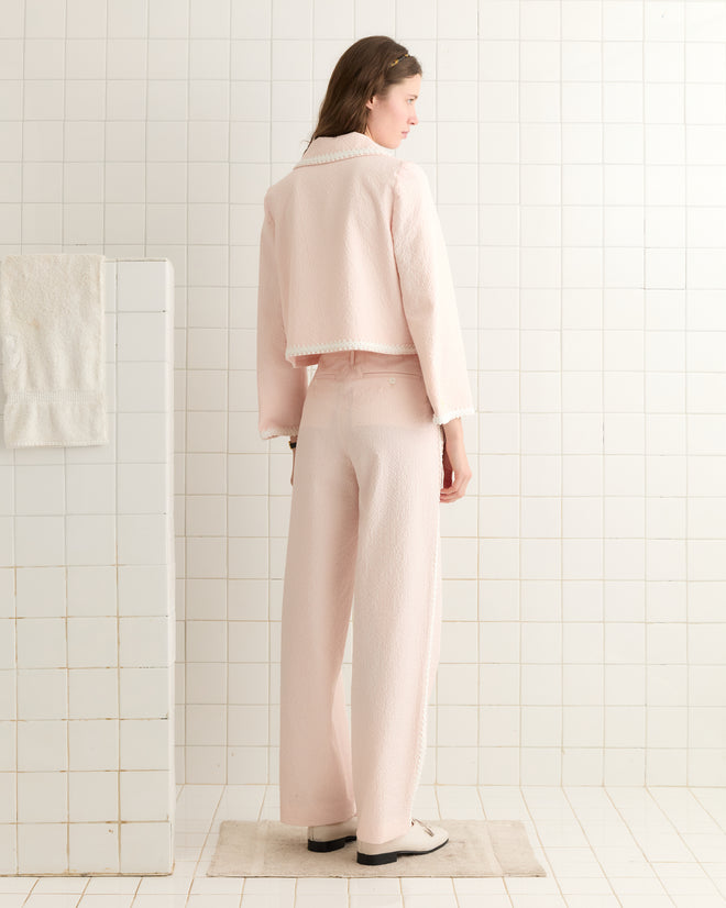 In a tiled bathroom, a person stands facing away wearing the Club Stripe Swing Jacket, a light pink outfit featuring antique pearl buttons inspired by 19th-century fashion.