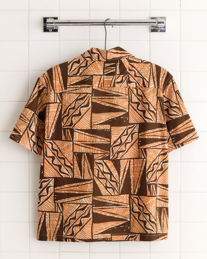 Cocoa Bean Shirt - S/M: Short-sleeved shirt with brown and orange geometric patterns in vintage barkcloth weave, displayed on a metal rack against a tiled wall.