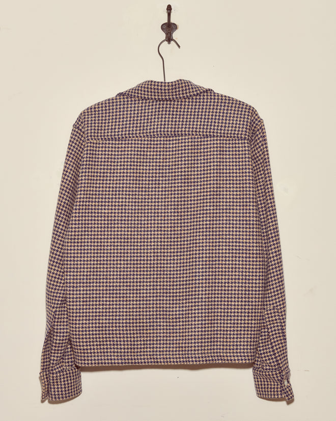 The Confetti Houndstooth Overshirt - S/M, with long sleeves, is displayed hanging on a hook from the back against a plain wall.