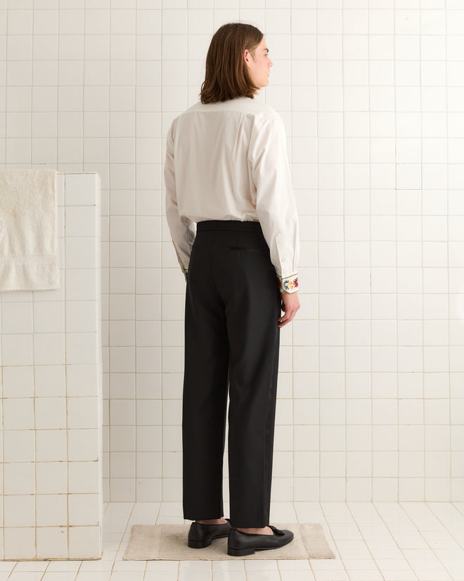 A person with long hair, wearing a white shirt and the stylish Connaught Smoking Trousers featuring a satin side stripe, stands in a tiled room facing away from the camera.