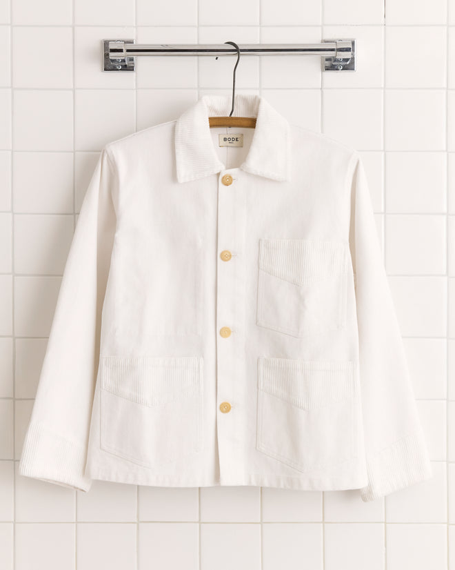 The Craft Jacket - White is a canvas workwear jacket featuring large front pockets and subtle corduroy trim, displayed on a hanger against a white tiled wall.