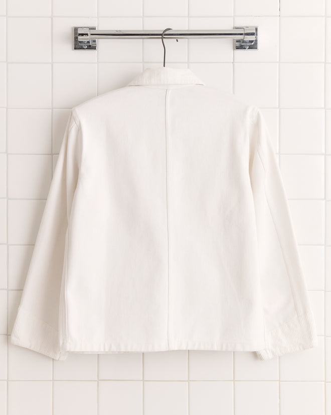 The Craft Jacket - White, made from heavyweight cotton twill with subtle corduroy trim, hangs on a hanger against a tiled white wall, displayed from the back.