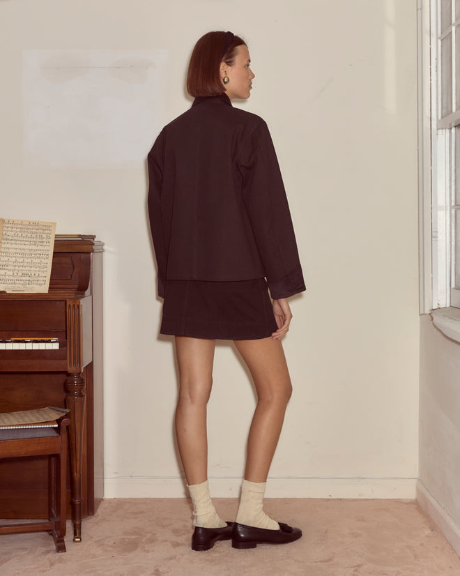 A woman wearing the Craft Jacket - Black, made from heavyweight cotton twill canvas, stands by a piano near a softly lit window, facing away.