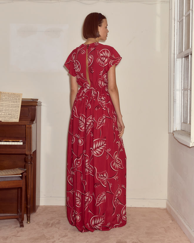 A woman in the Creeping Begonia Dress, featuring a red botanical print, stands near a piano by a window, facing away.