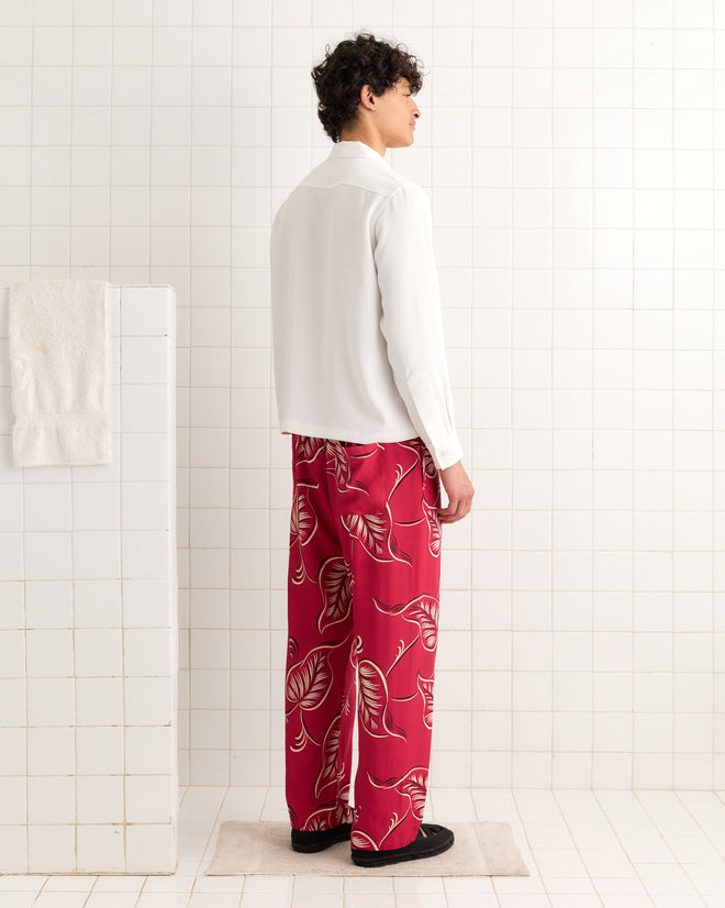 Man wearing Creeping Begonia Pajama Pants and a white shirt stands sideways in a white tiled bathroom.