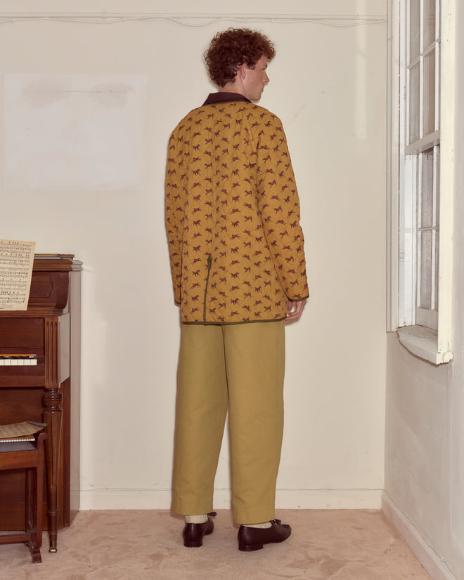 A person in the Crittendon Liner Coat with a yellow dog motif, knit collar, and tan pants stands by a piano and window facing the corner.