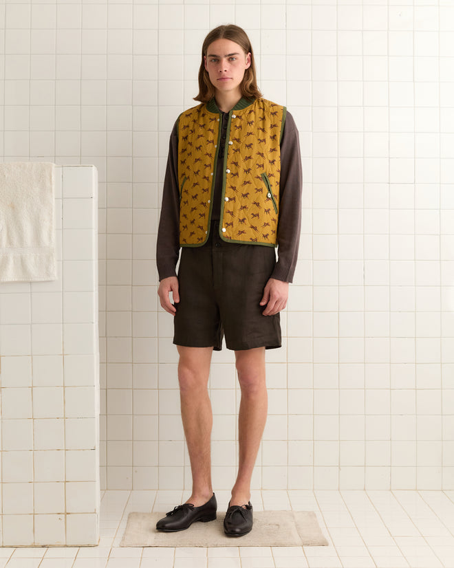 A person stands on a bath mat in a tiled room, wearing the Crittendon Quilted Vest in yellow with a dark shirt and shorts.