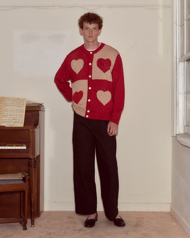 Wearing the Lamé Hearts Cardigan—a lightweight red knit with heart shapes inspired by Alice in Wonderland—a person stands by a piano in a softly lit room.
