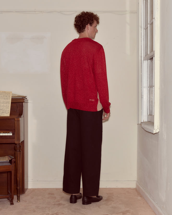 A man in a red Lamé Hearts Cardigan and black pants stands facing a wall near a window and an upright piano, his whimsical style echoing the playful spirit of Alice in Wonderland.