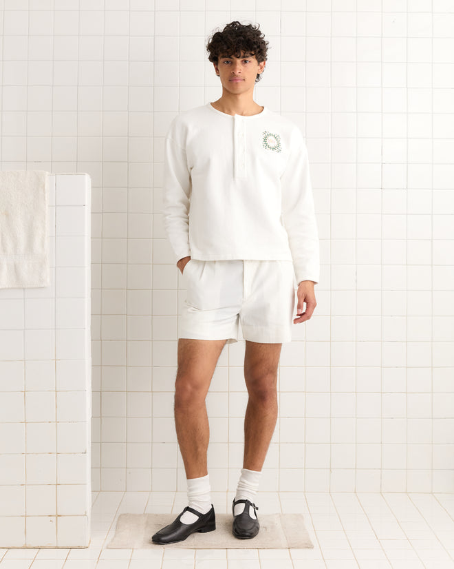 A young man wears the Daisy Never Tell Henley, a boxy fit, 100% cotton white long-sleeve shirt, paired with shorts, socks, and black shoes while standing in a tiled room.