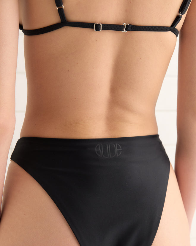 Someone wearing the Deco Bikini Bottoms - Black, shown from the back, against a white paneled background.