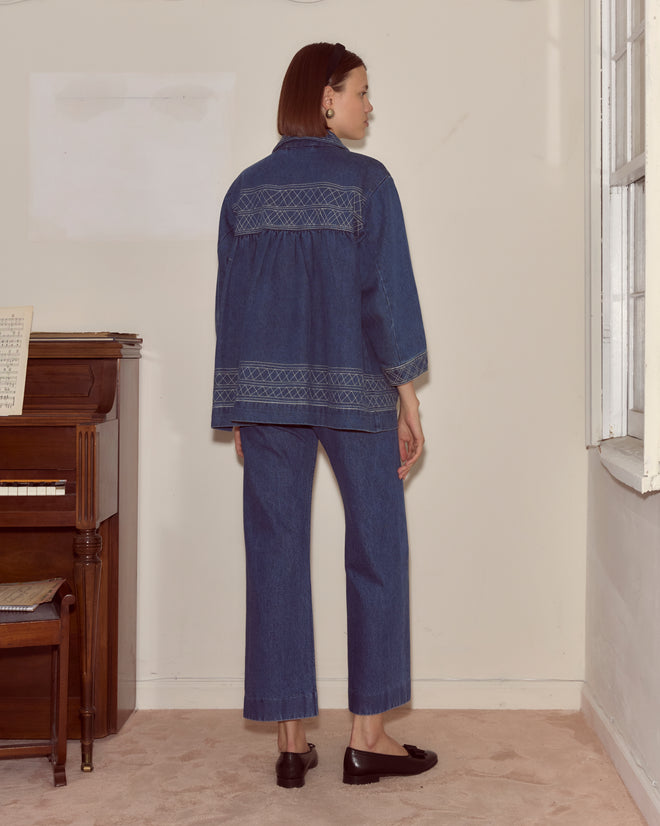 A woman in the Embroidered Denim Quincy Jacket and matching pants stands indoors by a window and piano, effortlessly channeling Bode-inspired style.