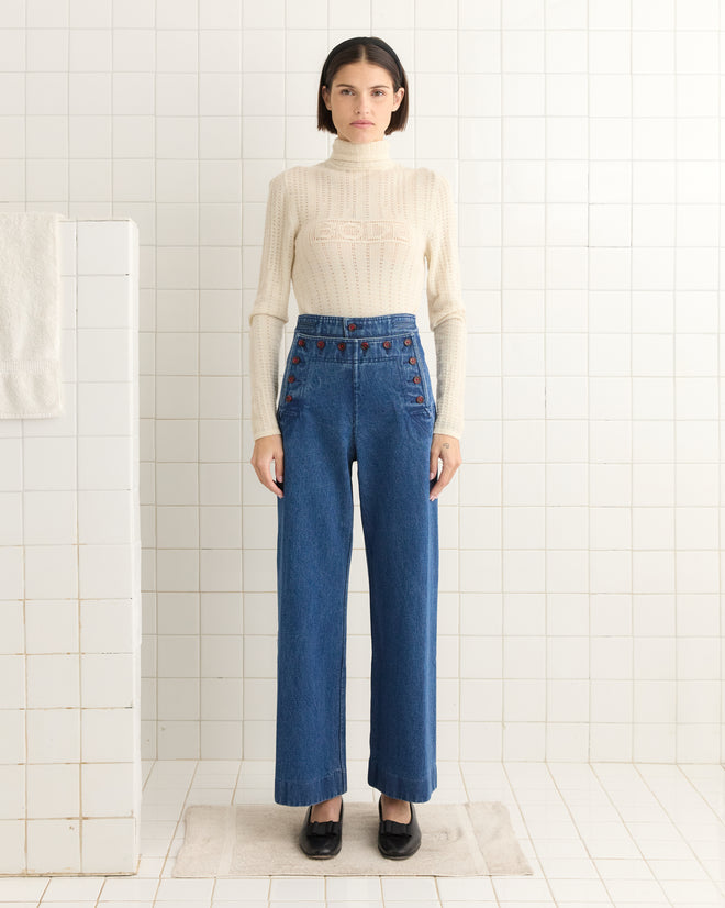 A person in a beige sweater and Denim Sailor Trousers stands on a bathroom mat against a tiled wall.