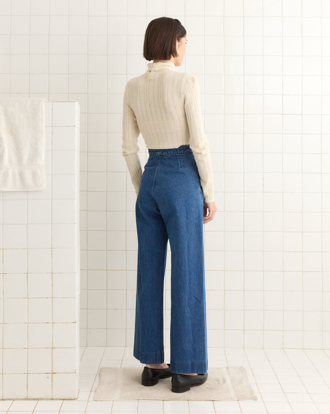 In a tiled bathroom, a person wearing a white turtleneck and Denim Sailor Trousers inspired by the classic 1940s style is facing away.