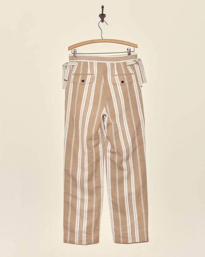 Desert Stripe Trousers in tan and white vertical stripes, displayed on a wooden hanger against a plain off-white wall.
