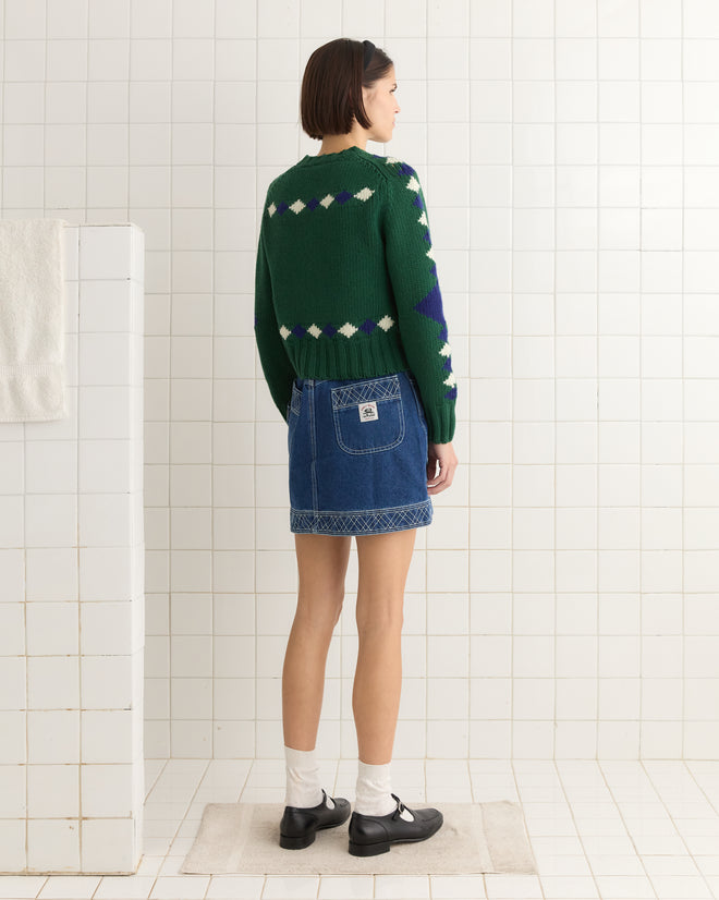 A person wearing a green Diamond Corral Sweater and a denim skirt stands on a white tiled bathroom floor.