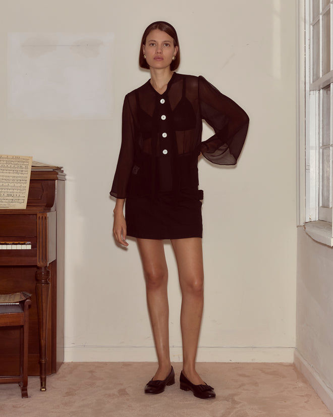 A woman wearing the Dottie Blouse, a sheer silk top with antique pearl buttons, stands indoors next to a piano with sheet music.