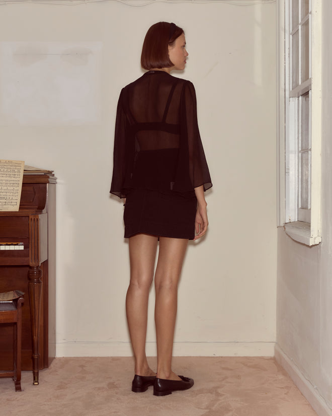 A woman in the Dottie Blouse—100% silk, sheer, with antique pearl buttons—and a skirt stands by a window and a piano.