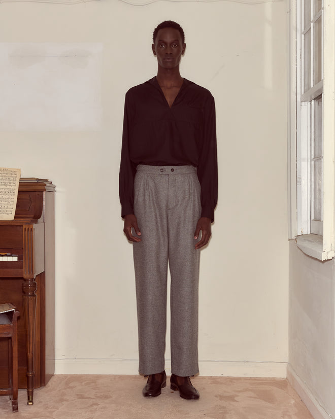 A person stands indoors by a piano, dressed in a black shirt and classic Dovecote Trousers, gazing directly ahead.
