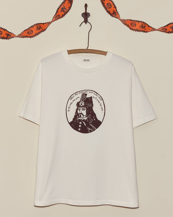 The Dracula Tee features a circular black illustration of a historical figure on a white shirt, hanging on a hook with festive garland above—ideal as a vintage or unique Halloween tee.
