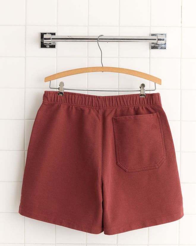 Burgundy Pennant Shorts made from 100% cotton with an elastic waistband and back pocket hang on a wooden hanger against a white tiled wall.