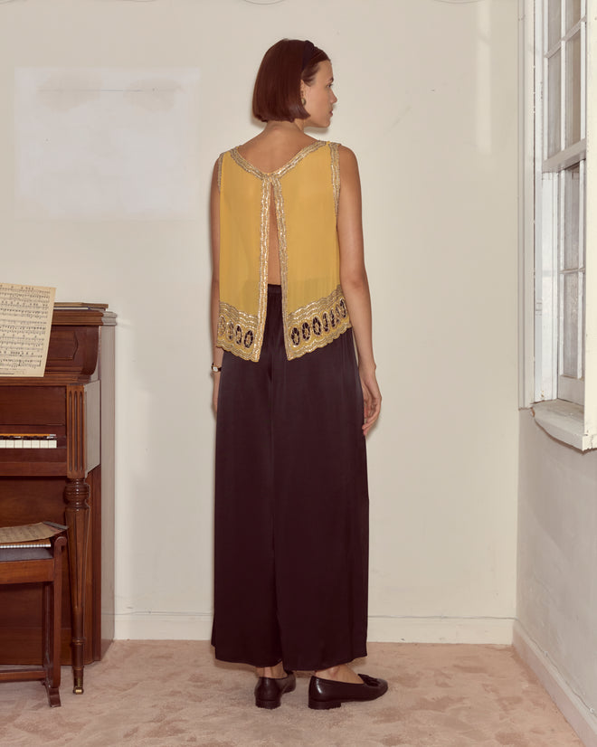 A woman in a yellow Ducat Tank and black pants stands by a piano, facing away, near a window.