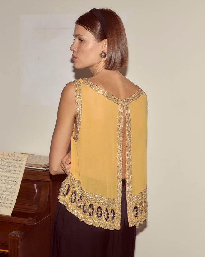 A woman with short hair, wearing the yellow Ducat Tank, stands by a piano with sheet music, facing away from the camera.