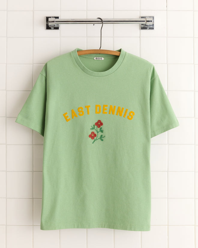 The East Dennis Tee, featuring vibrant red floral embroidery and made from 100% cotton, hangs gracefully on a wooden hanger against a tiled wall.