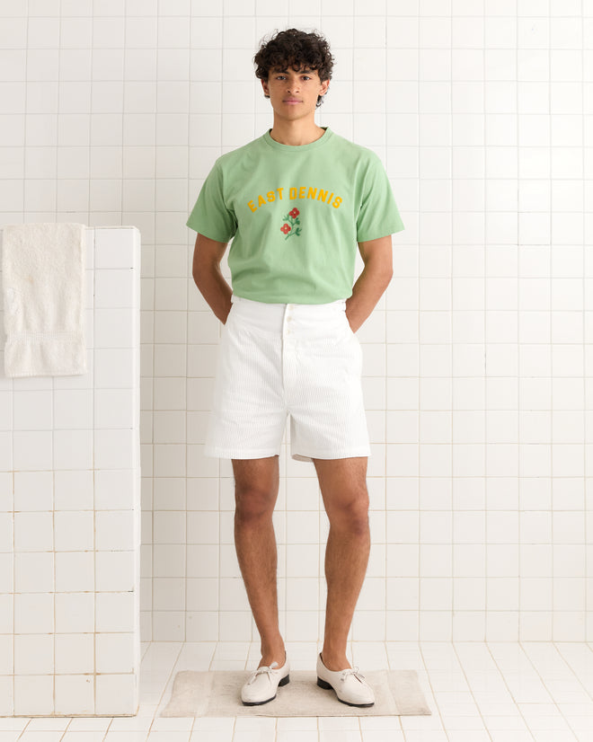 A person stands in a tiled room wearing a green East Dennis Tee made of 100% cotton, paired with white shorts and loafers.