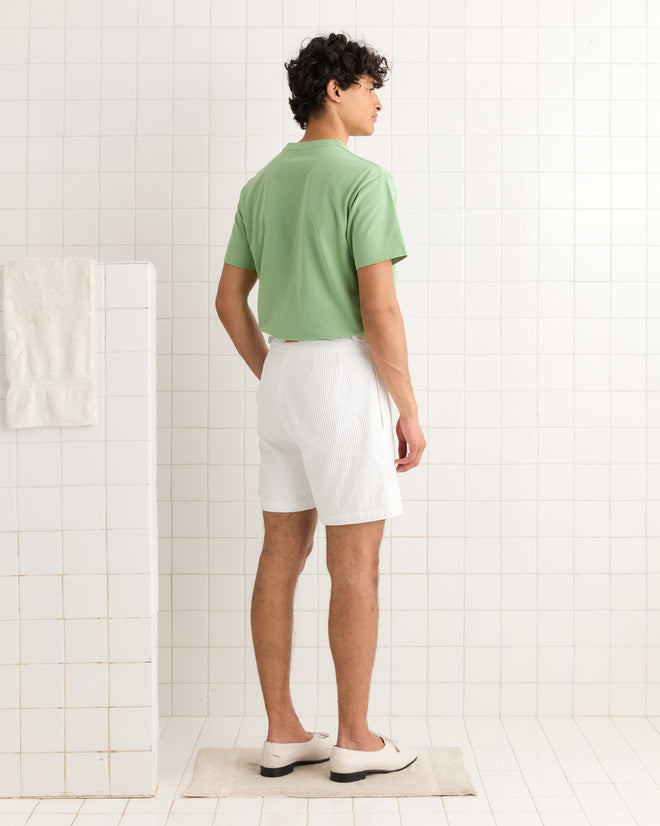 Man standing in a tiled room, facing away, wearing the green East Dennis Tee, white shorts, and white shoes.