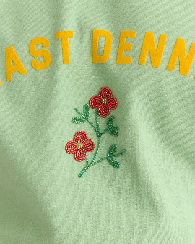 The East Dennis Tee is a green 100% cotton shirt featuring yellow text and three red beaded flowers with green stems embroidered below, perfect for those who love graphic tees inspired by East Dennis style.