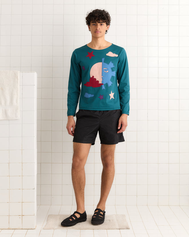 A man in an Eclipse Long Sleeve Shirt with a sun, moon, and stars design and black shorts stands on a bath mat in a white tiled bathroom.