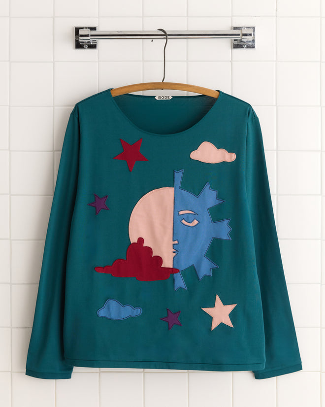 Displayed on a tiled wall, the Eclipse Long Sleeve Shirt in teal showcases a stylized sun, moon, and stars with clouds. The retro design exudes a 1960s vibe with its distinctive artistic flair.