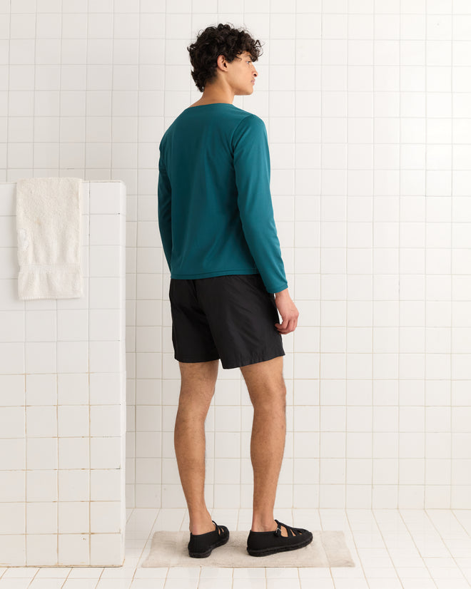 A man in the Eclipse Long Sleeve Shirt with stylized sun, moon, and stars and black shorts stands on a bath mat in a tiled bathroom, facing away.