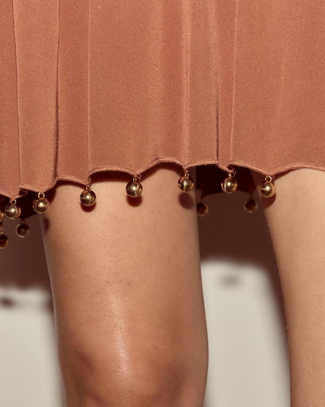 The Eddy Dress features a brown skirt hem adorned with gold ball ornaments and a chic drop-waist silhouette, creating an elegant couture-inspired look.