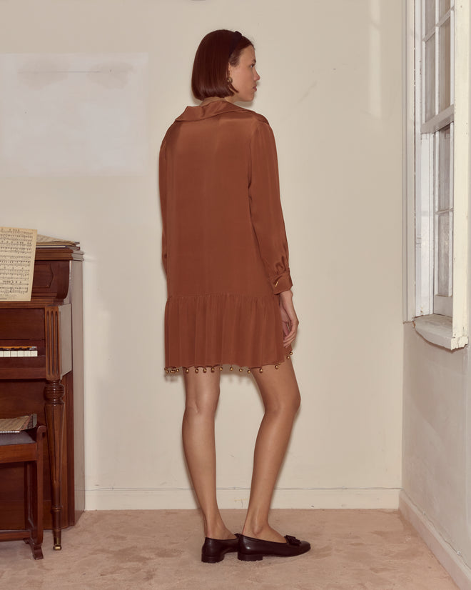 A woman wearing the Eddy Dress in brown silk stands indoors by a window and piano, facing away from the camera.