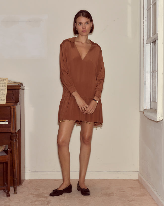 A woman in the Eddy Dress, a 100% silk brown dress with a drop-waist silhouette, stands indoors by a window and piano, looking at the camera.