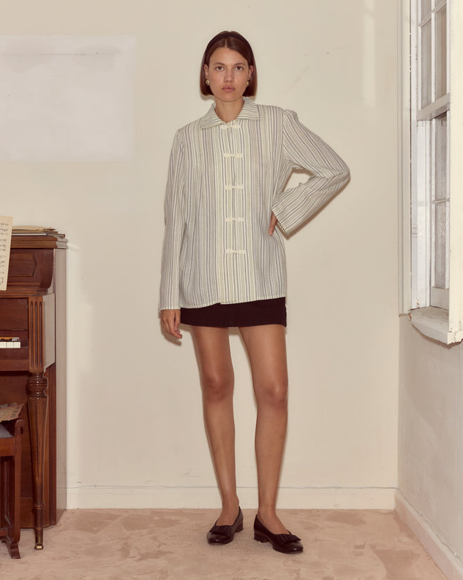 Woman wearing the Electric Light Blouse, a pale yellow striped top with ivory frog closures, paired with a black skirt and black shoes, stands indoors by a piano and window.